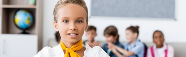 Panoramic crop of schoolgirl looking at camera with miltiethnic classmates at background — Stock Photo