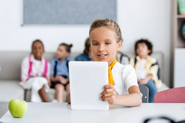Selective focus of schoolgirl using digital tablet near apple and multicultural friends at background in classroom — Stock Photo