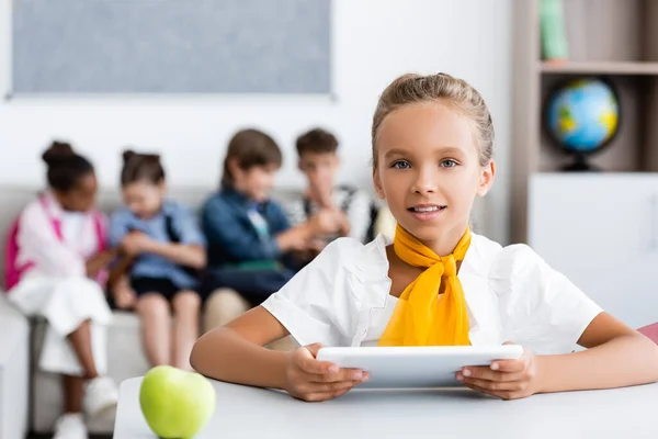 Enfoque selectivo de colegiala sosteniendo tableta digital cerca de manzana en el escritorio y amigos multiétnicos en el aula - foto de stock