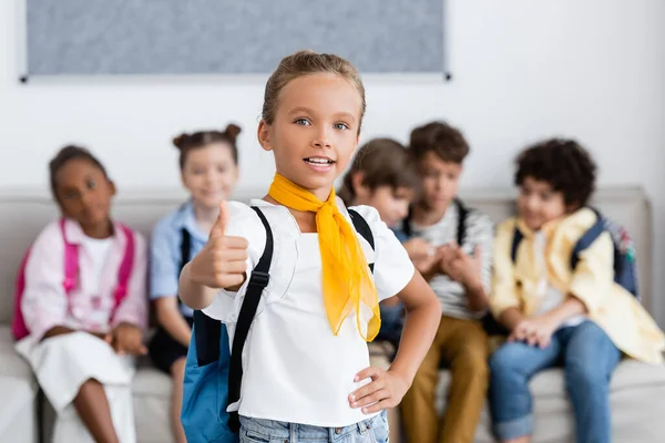 Selective focus of schoolgirl with backpack showing like gesture near multiethnic friends in school — Stock Photo