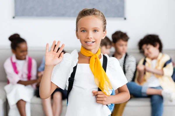 Selektiver Fokus einer Schülerin mit Rucksack, die mit multikulturellen Freunden im Hintergrund in die Kamera winkt — Stockfoto