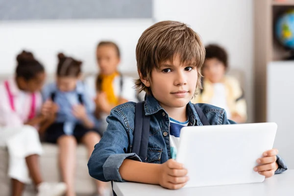 Enfoque selectivo del escolar sosteniendo la tableta digital y mirando la cámara en el escritorio en el aula - foto de stock