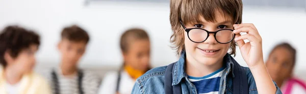 Concepto panorámico de colegial sosteniendo anteojos y mirando a la cámara en la escuela - foto de stock