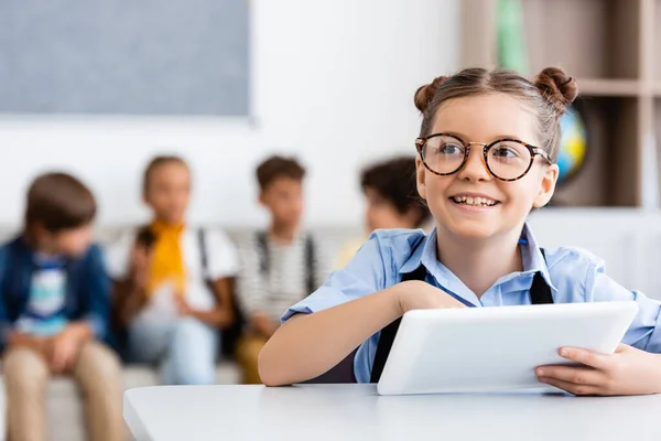 Enfoque selectivo de la colegiala con tableta digital sentada en el escritorio cerca de amigos en el fondo en el aula - foto de stock