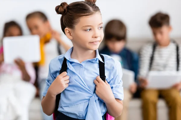 Enfoque selectivo de colegiala sosteniendo mochila cerca de amigos multiétnicos en el fondo - foto de stock