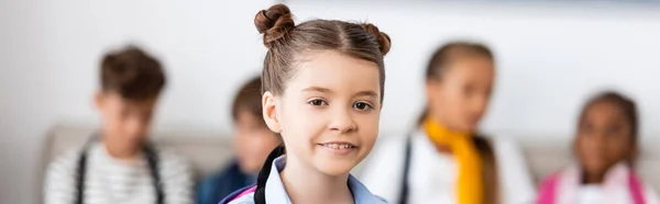 Panoramic shot of schoolgirl looking at camera near multicultural classmates in school — Stock Photo