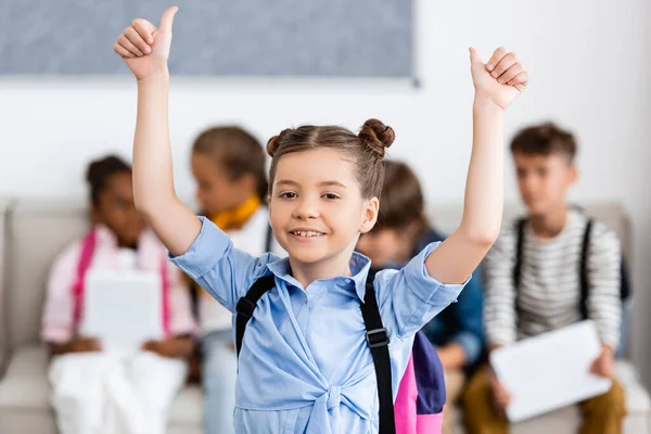 Selective focus o schoolgirl with backpack showing thumbs up near multiethnic friends at background — Stock Photo