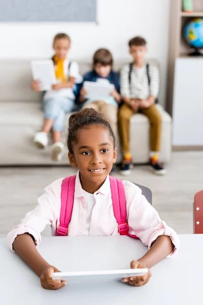 Enfoque selectivo de la colegiala afroamericana con mochila sosteniendo tableta digital en el escritorio cerca de amigos en el aula - foto de stock