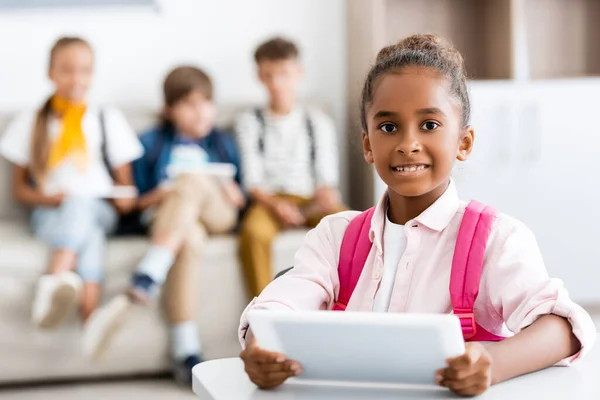 Foco seletivo da menina americana africana segurando tablet digital na mesa com colegas de classe em segundo plano na escola — Fotografia de Stock