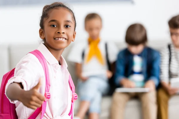 Selective focus of african american schoolgirl with backpack showing like at camera in classroom — Stock Photo