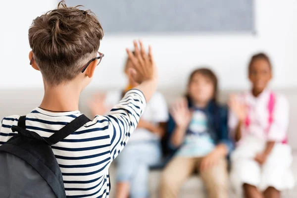 Selective focus of schoolboy with backpack waving hand at multiethnic classmates — Stock Photo