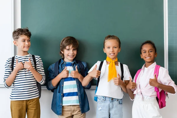 Des écoliers multiculturels montrant leurs pouces près du tableau noir à l'école — Photo de stock