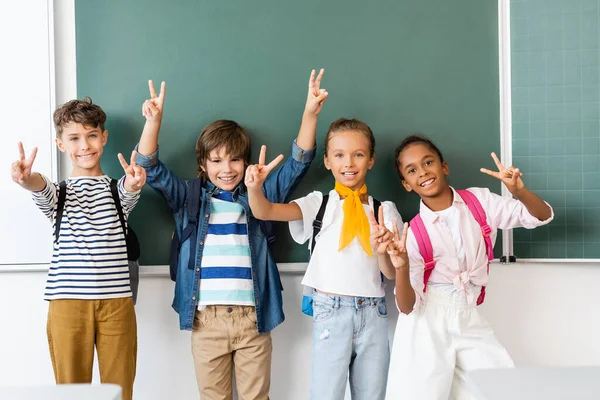 Selektiver Fokus multiethnischer Schüler, die in der Nähe der Tafel im Klassenzimmer vor der Kamera das Siegeszeichen zeigen — Stockfoto