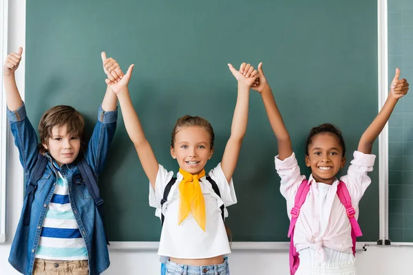 Multiethnic schoolkids showing approval gesture near chalkboard — Stock Photo