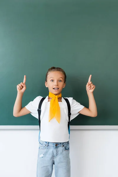Écolière excitée avec sac à dos pointant avec les doigts au tableau noir — Photo de stock