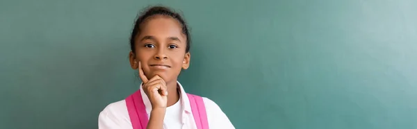 Panoramic shot of african american schoolgirl with finger near cheek looking at camera near chalkboard — Stock Photo