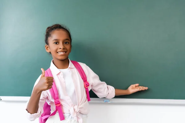 Selective focus of african american schoolgirl showing like gesture and pointing with hand at chalkboard — Stock Photo