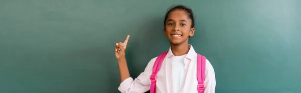 Horizontal concept of african american schoolgirl pointing at green chalkboard in classroom — Stock Photo