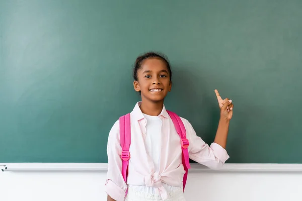 African american schoolchild with backpack looking at camera while pointing with finger near chalkboard — Stock Photo