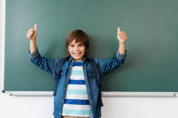 Schoolboy with backpack showing thumbs up near green chalkboard — Stock Photo