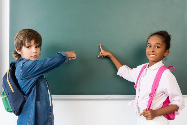 Multicultural schoolkids looking at camera while pointing at chalkboard in classroom — Stock Photo