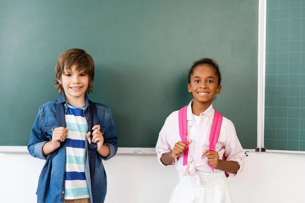 Schoolboy with backpack standing near african american friend and chalkboard in classroom — Stock Photo