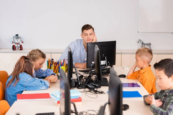 Selective focus of teacher looking at multiethnic pupils near computers in stem school — Stock Photo