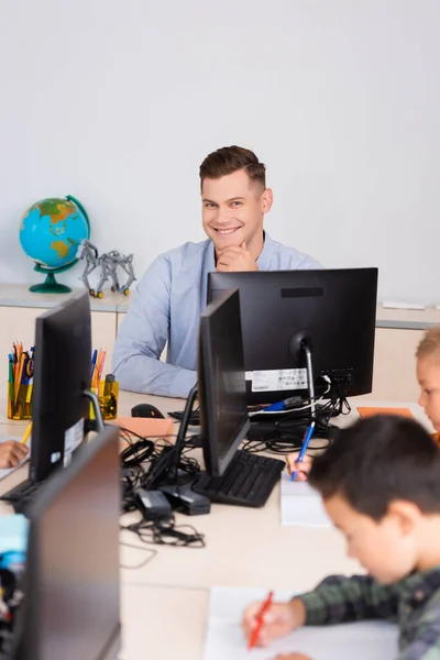 Selektiver Fokus des Lehrers auf die Kamera in der Nähe von Computern und multiethnischen Schülern im Klassenzimmer — Stockfoto