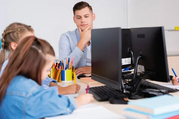 Foco seletivo de professor sentado perto de alunos que escrevem em cadernos perto de computadores na escola — Fotografia de Stock