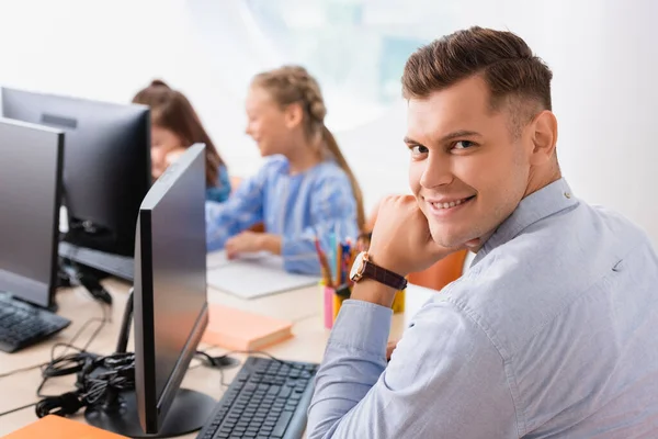 Selective focus of teacher looking at camera near computers and pupils in classroom — Stock Photo
