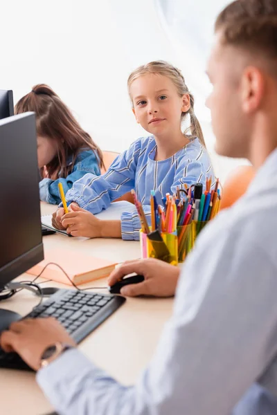 Foco seletivo de estudante com caneta olhando para professor perto de computadores em sala de aula — Fotografia de Stock