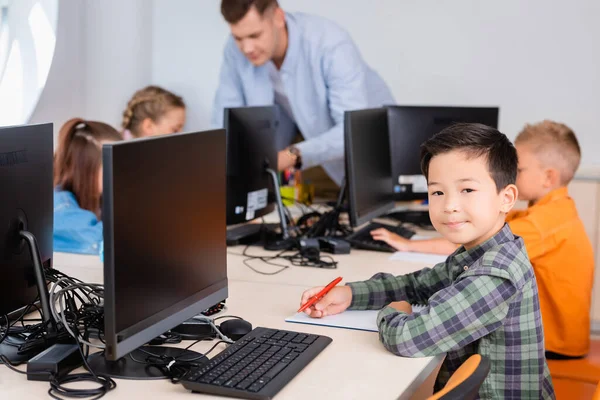 Foyer sélectif de asiatique écolier regarder caméra tout en écrivant sur notebook près des ordinateurs dans tige école — Photo de stock