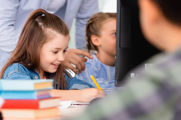 Enfoque selectivo del profesor de pie cerca de la colegiala escribiendo en el cuaderno en la escuela madre - foto de stock