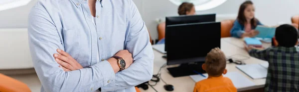 Panoramic crop of teacher with crossed arms standing near pupils and computers in stem school — Stock Photo