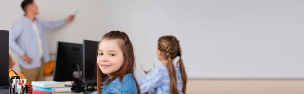 Горизонтальне зображення школярки, яка дивиться на камеру під час уроку поблизу комп'ютерів у стовбуровій школі — стокове фото