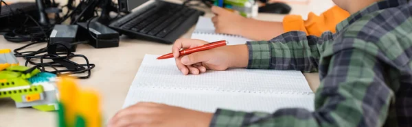 Panoramic crop of schoolboy writing on notebook near computer in school — Stock Photo
