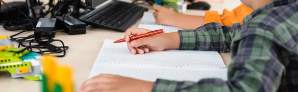 La cosecha panorámica del colegial que escribe en el cuaderno cerca del ordenador en la escuela - foto de stock