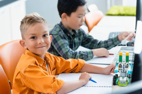 Selective focus of schoolboy looking at camera near robot and asian friend in stem school — Stock Photo