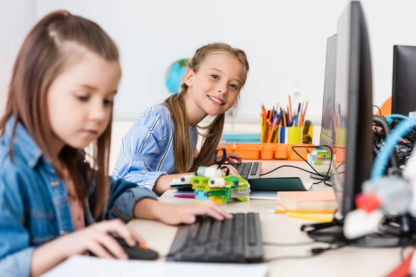 Selective focus of schoolgirl looking at camera near robot and computers during lesson in stem school — Stock Photo