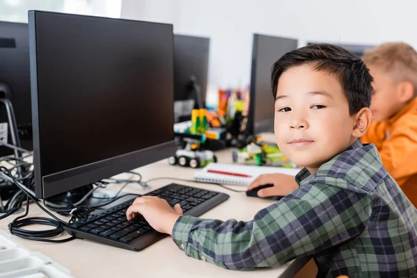 Enfoque selectivo de asiático colegial mirando cámara mientras usando computadora en aula - foto de stock