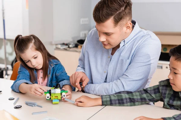 Selective focus of teacher pointing at robot with parts near multiethnic kids in classroom — Stock Photo