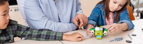 Panoramic shot of teacher pointing at robot with parts near multicultural schoolchildren in classroom — Stock Photo