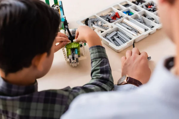 Back view of teacher sitting near asian schoolboy modeling robot at table in stem school — Stock Photo