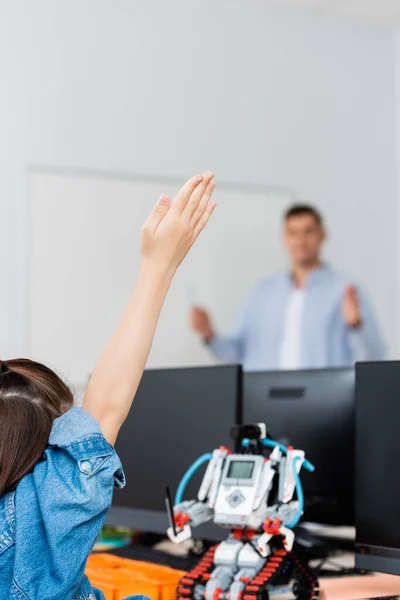 Selective focus of schoolgirl with raised hand sitting near robot and computers in classroom — Stock Photo