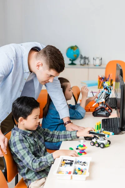 Selective focus of teacher standing near multiethnic pupils modeling robots in classroom — Stock Photo