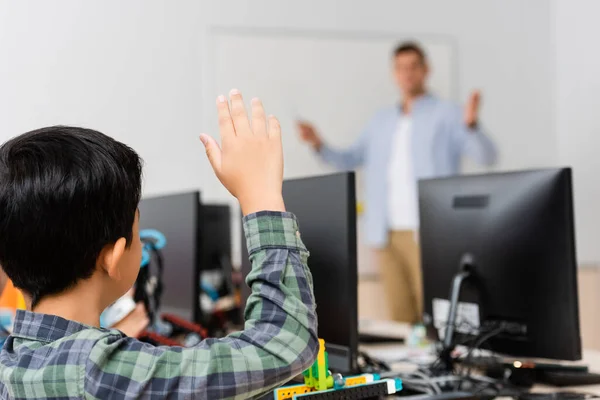 Selective focus of schoolboy with raised hand sitting near robot and computers during lesson in stem school — Stock Photo