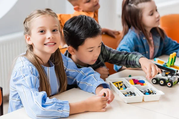 Selective focus of schoolgirl looking at camera near multiethnic friends modeling robot in classroom — Stock Photo