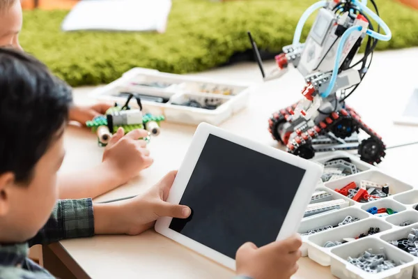 Selective focus of multiethnic pupils using digital tablet while constructing robot in classroom — Stock Photo