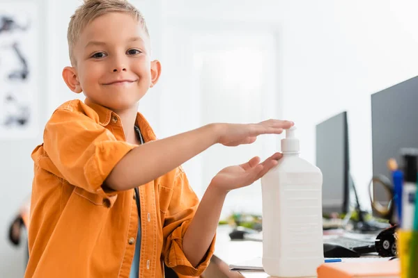 Selective focus of schoolboy using hand sanitizer and looking at camera in classroom — Stock Photo