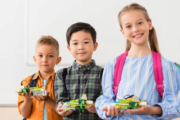 Enfoque selectivo de alumnos multiétnicos sosteniendo robots y mirando a la cámara en la escuela de tallos — Stock Photo